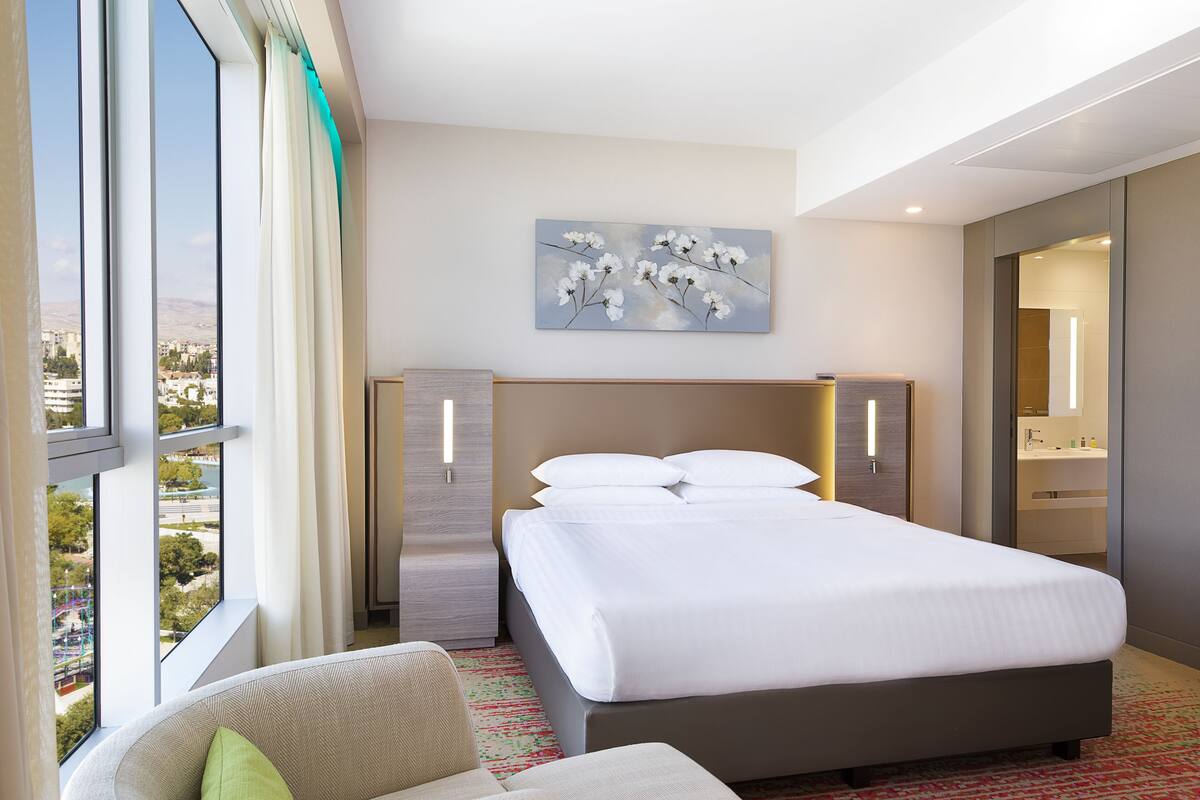 A hotel room with a large bed, a view of the city, a painting on the wall, and a bathroom visible through an open door.