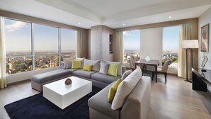 A spacious living room with a large sectional sofa, a coffee table, and a dining area with a table and chairs. The room has large windows offering a city view.