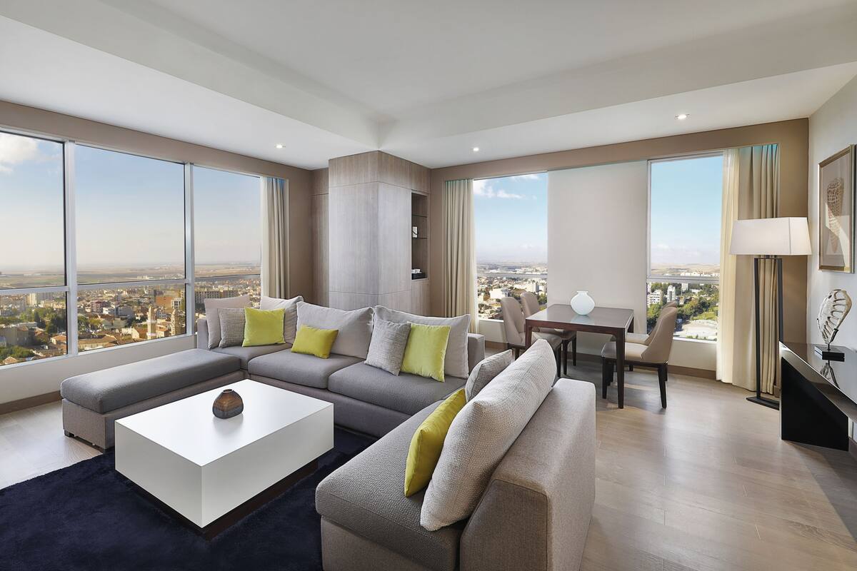 A spacious living room with a large sectional sofa, a coffee table, and a dining area with a table and chairs. The room has large windows offering a city view.