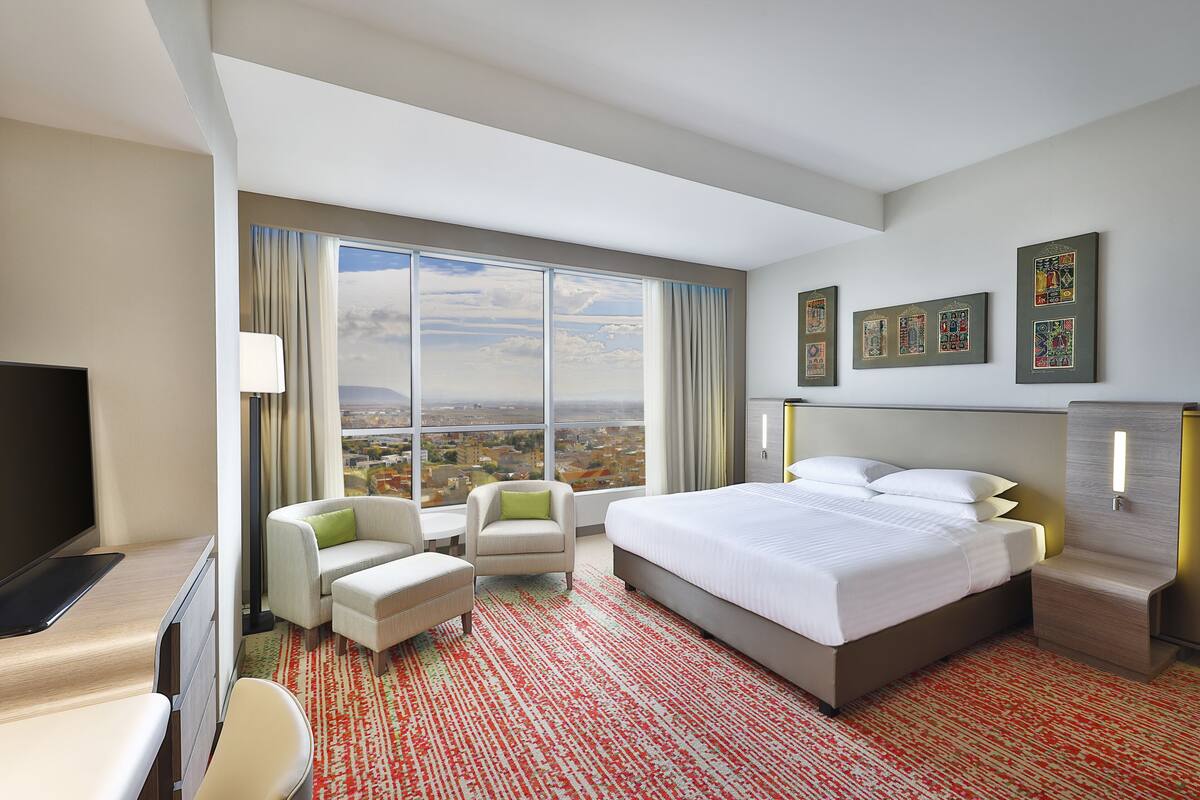 A modern hotel room with a large bed, a desk, a chair, and a view of the city.