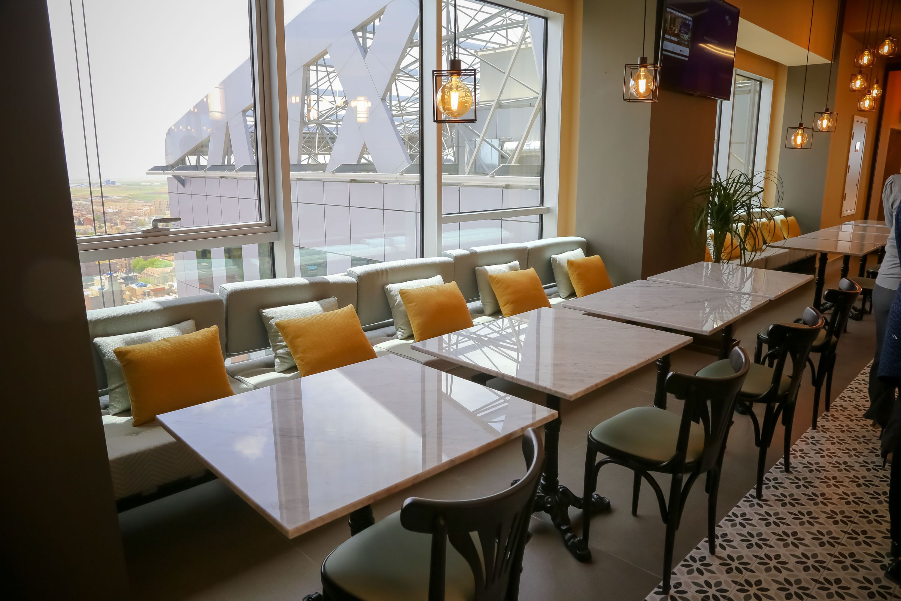 A modern restaurant with high tables and chairs, large windows with a view of a building, and hanging pendant lights.