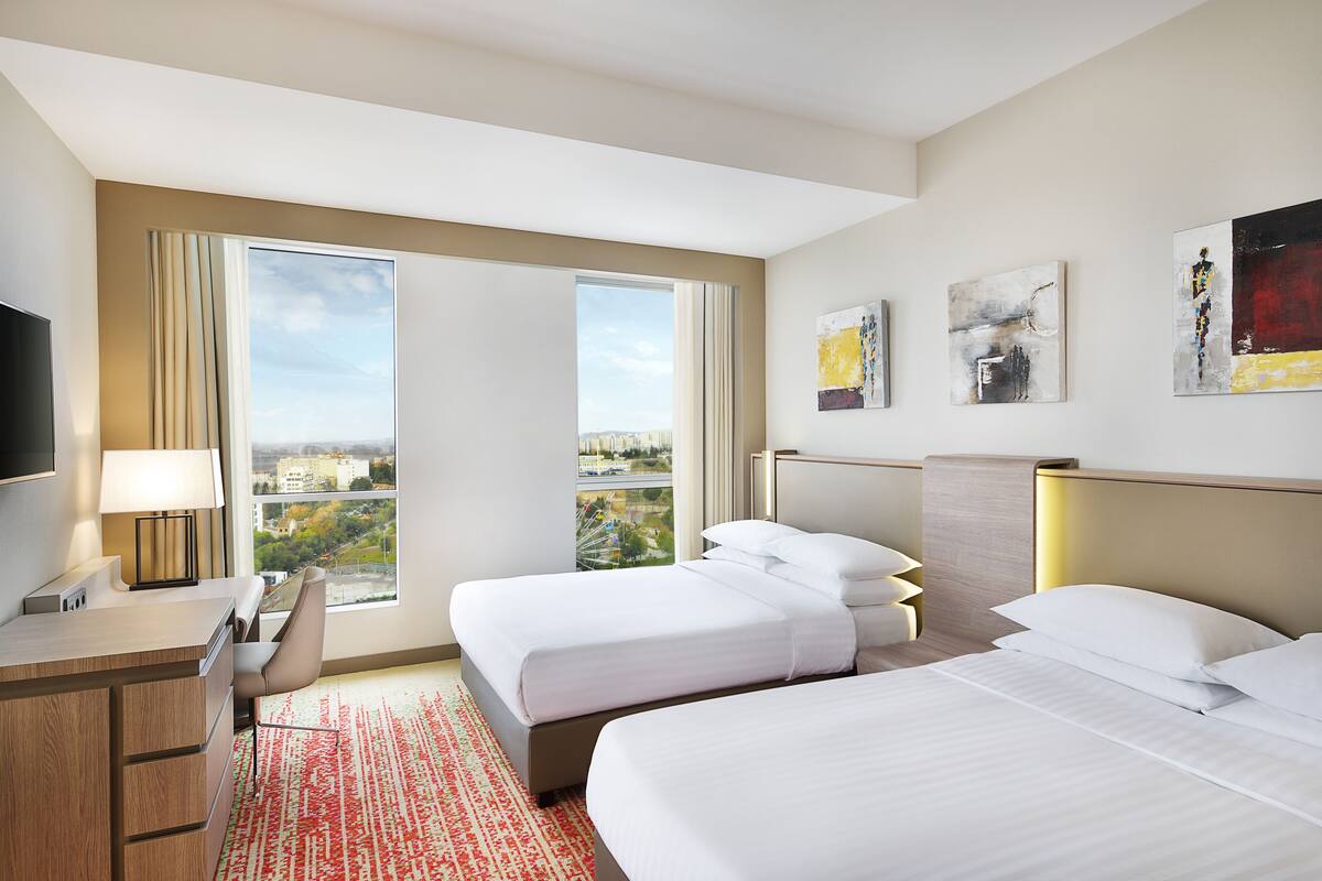 A hotel room with two beds, a desk, a chair, and a large window offering a city view.