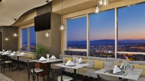A restaurant with tables set for dining, large windows with a city view, and a flat-screen TV mounted on the wall.