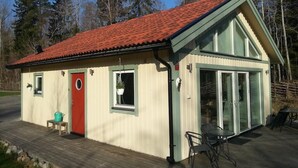 Un petit bâtiment d’un seul étage, avec un toit recouvert de tuiles rouges, une porte rouge et une porte vitrée donnant sur un espace extérieur abrité. Il y a un petit coin salon extérieur avec une table et des chaises.