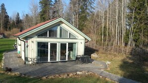 Un petit bâtiment d’un seul étage, doté d’un toit en pente et de grandes portes vitrées, situé sur une terrasse dallée avec des espaces assise extérieurs.