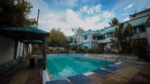A swimming pool with a tiled deck, surrounded by buildings with blue and white exteriors, and some greenery. - Seashell Beach Resort (Dahanu)