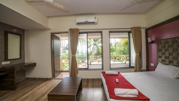 A hotel room with a bed, a wooden bench, a TV, and a balcony with a view of palm trees. - Seashell Beach Resort (Dahanu)