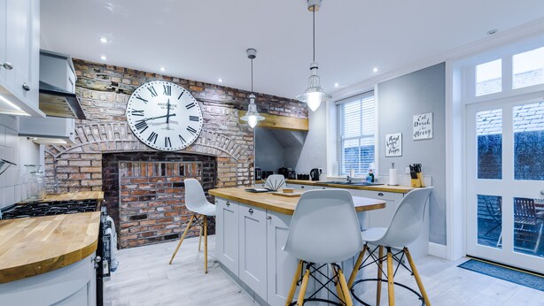 A modern kitchen with a brick wall, a large clock, and a bar area with stools. - Augustus House (Chester)