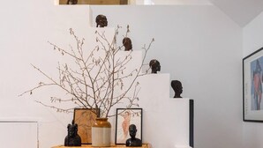 Ein Zimmer mit weißer Wand, einem hölzernen Regal mit Skulpturen, einer Vase mit Zweigen und gerahmten Kunstwerken.