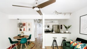 Un salon moderne avec un canapé vert, un coin repas avec une table ronde et des chaises, une cuisine équipée d’appareils électroménagers en acier inoxydable et un ventilateur de plafond.