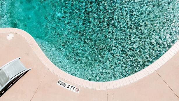 Une piscine d’un bleu éclatant, agrémentée d’un panneau indiquant « INTERDIT DE PLONGER – PROFONDEUR MINIMALE : 1,5 MÈTRE », d’une chaise longue et d’une terrasse carrelée.