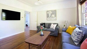 A living room with a flat-screen TV mounted on the wall, a grey sofa with yellow and patterned cushions, a wooden coffee table with a vase of flowers, and a framed picture on the wall. - Rodova Cottage with fireplace and enclosed backyard (Katoomba)