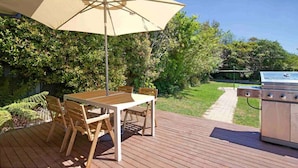 A wooden deck with a table and chairs under a large umbrella, a grill, and a pathway leading to a garden. - Rodova Cottage with fireplace and enclosed backyard (Katoomba)