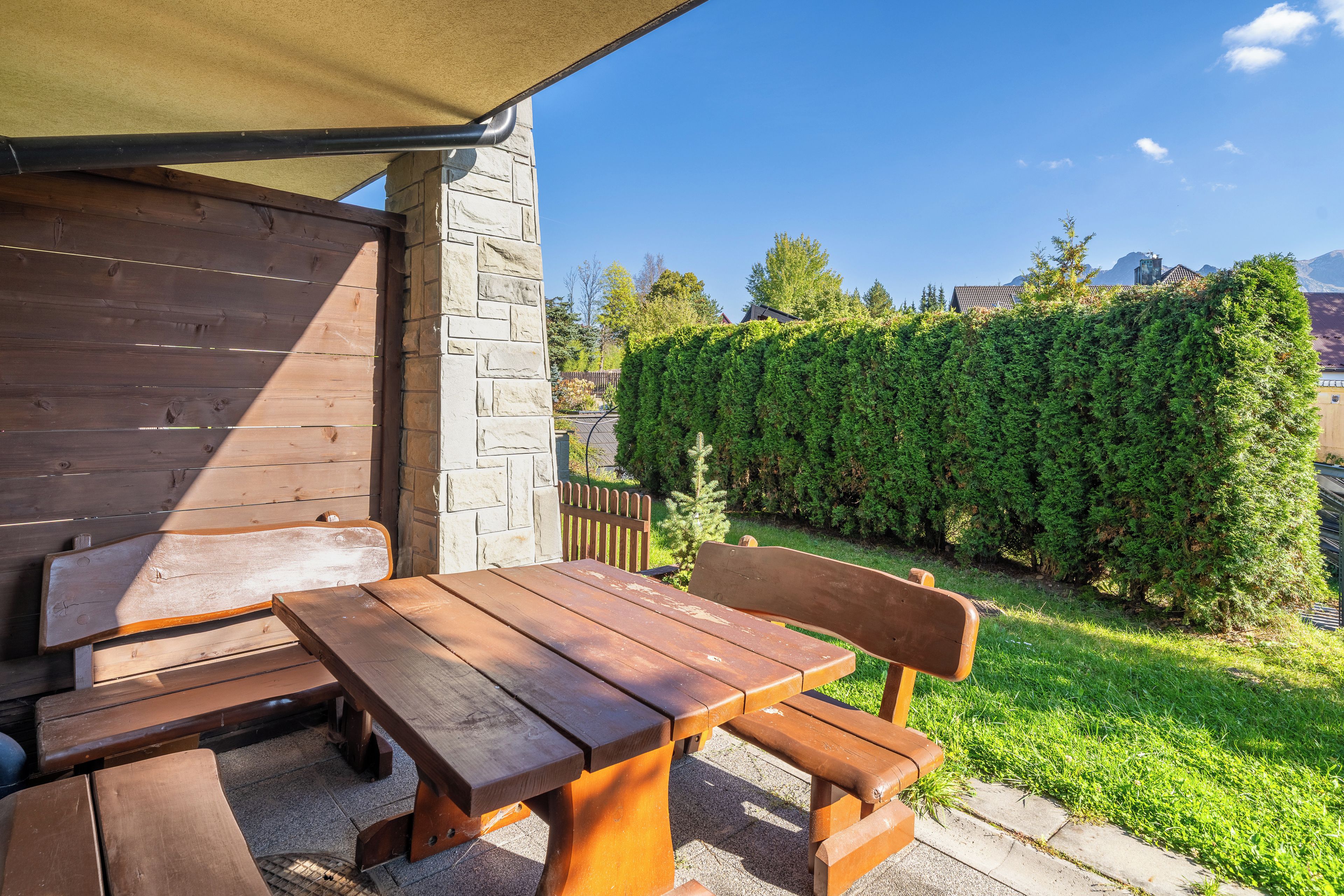 Una mesa de madera y bancos al aire libre, bajo una zona con sombra, con un muro de piedra y un seto al fondo.