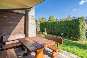 Une table et des bancs en bois, installés à l’extérieur sous un espace ombragé, avec un mur de pierres et une haie en arrière-plan.