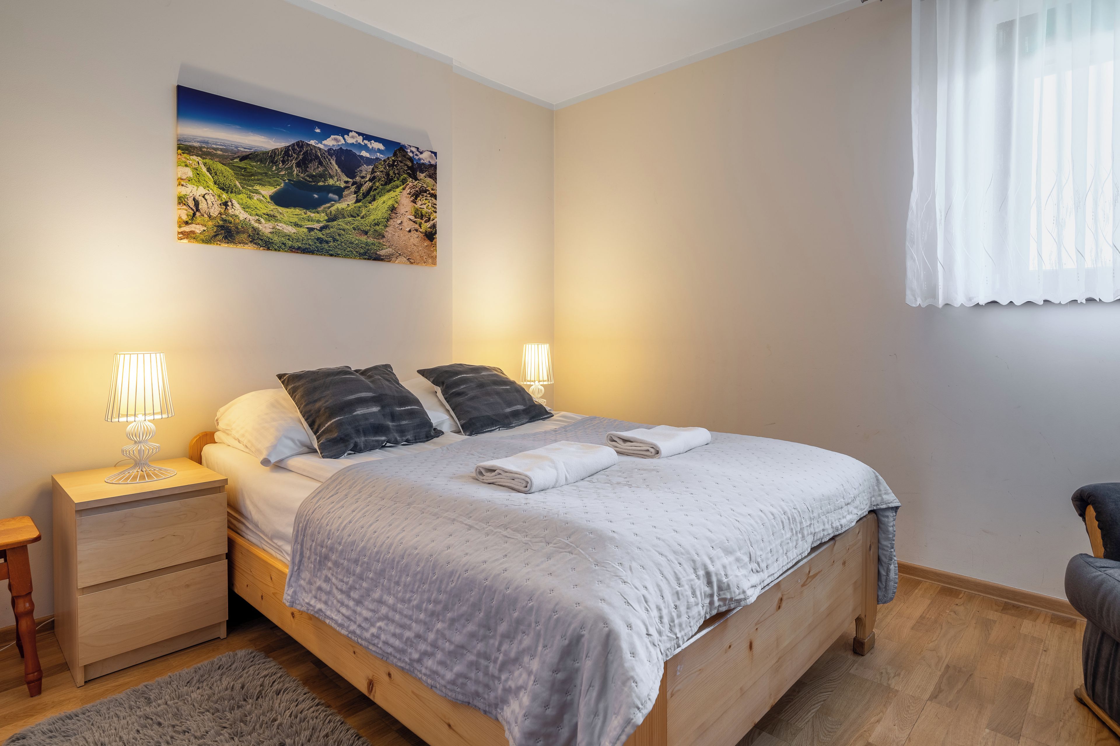 A bedroom with a wooden bed, two bedside lamps, a nightstand, a chair, a window with curtains, and a landscape painting on the wall.
