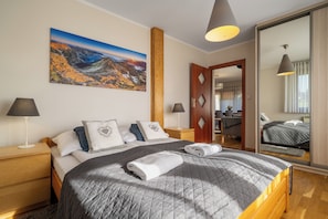 Une chambre à coucher avec un grand lit, deux tables de chevet, une commode, une porte en bois, un miroir et un tableau représentant un paysage de montagne.