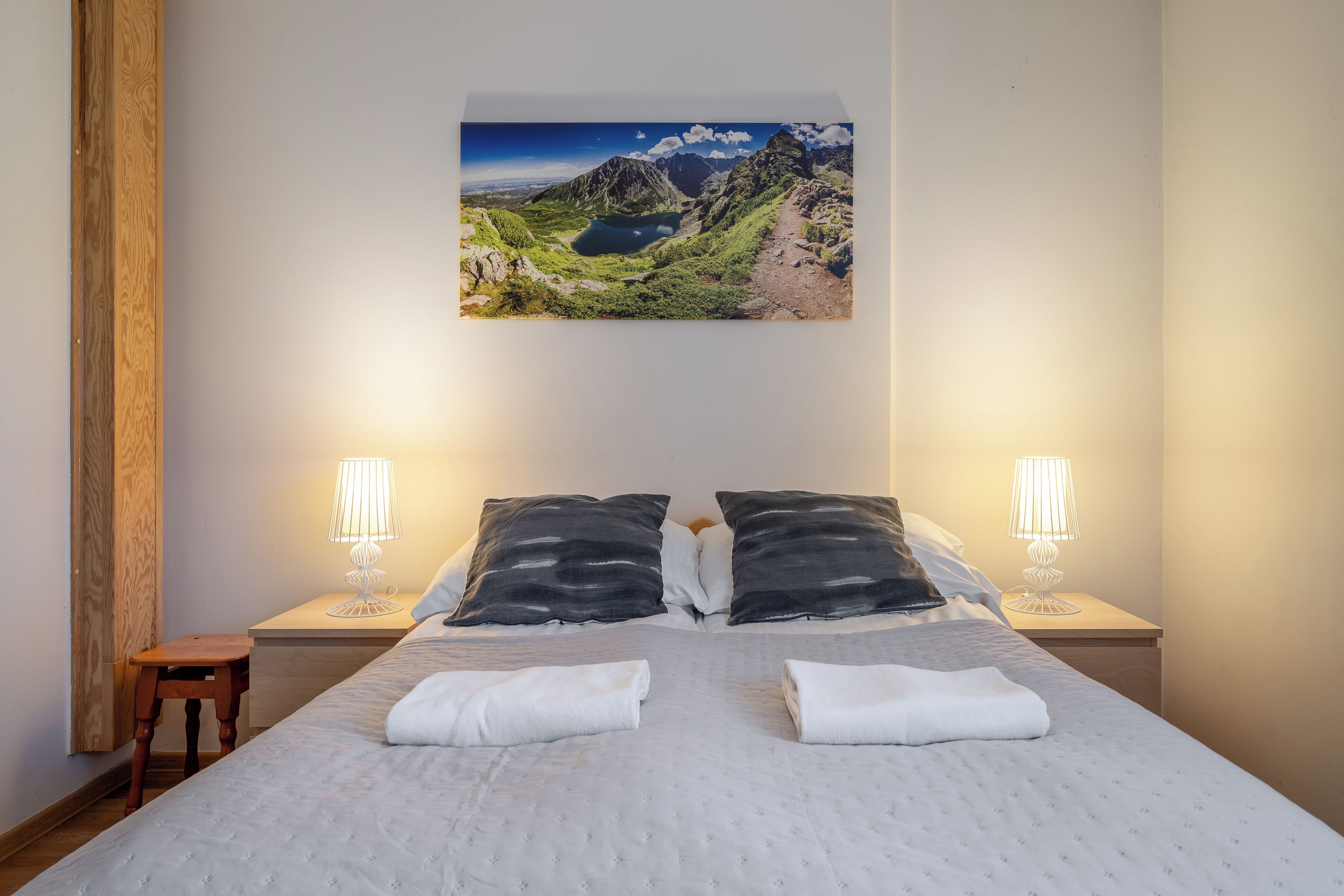 A bedroom with a large bed, two bedside lamps, a nightstand, a wooden chair, and a mountain landscape painting on the wall.
