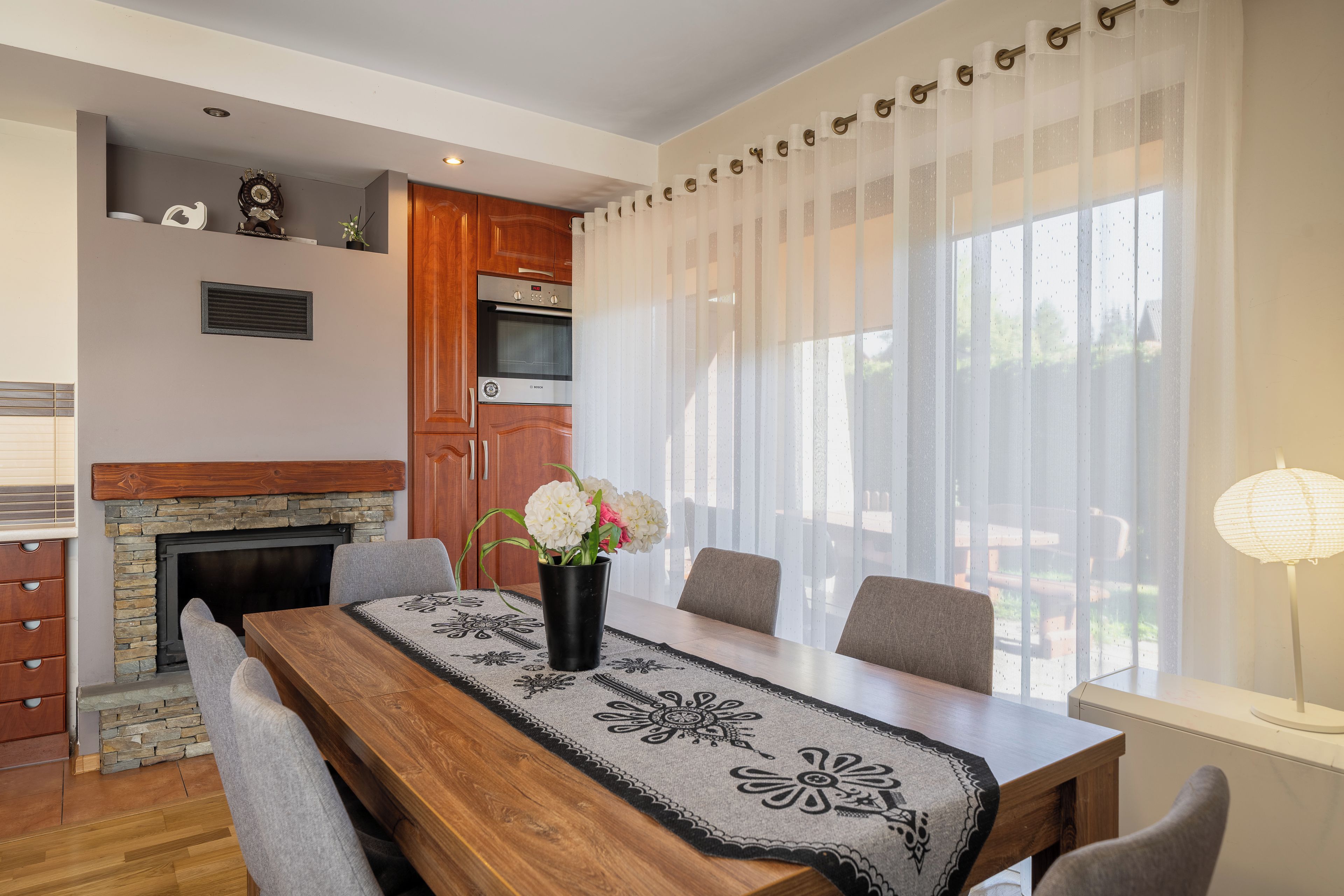 A dining area with a wooden table, chairs, a vase with flowers, a fireplace, and a window with sheer curtains.