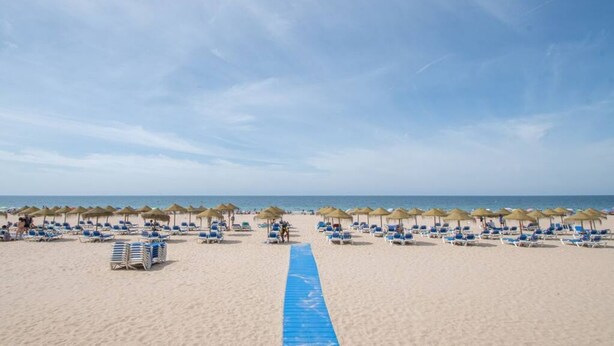 A beach with rows of sun loungers and thatched umbrellas, a clear sky, and a blue pathway leading to the sea. - El pentagrama de Cadiz (Cádiz)