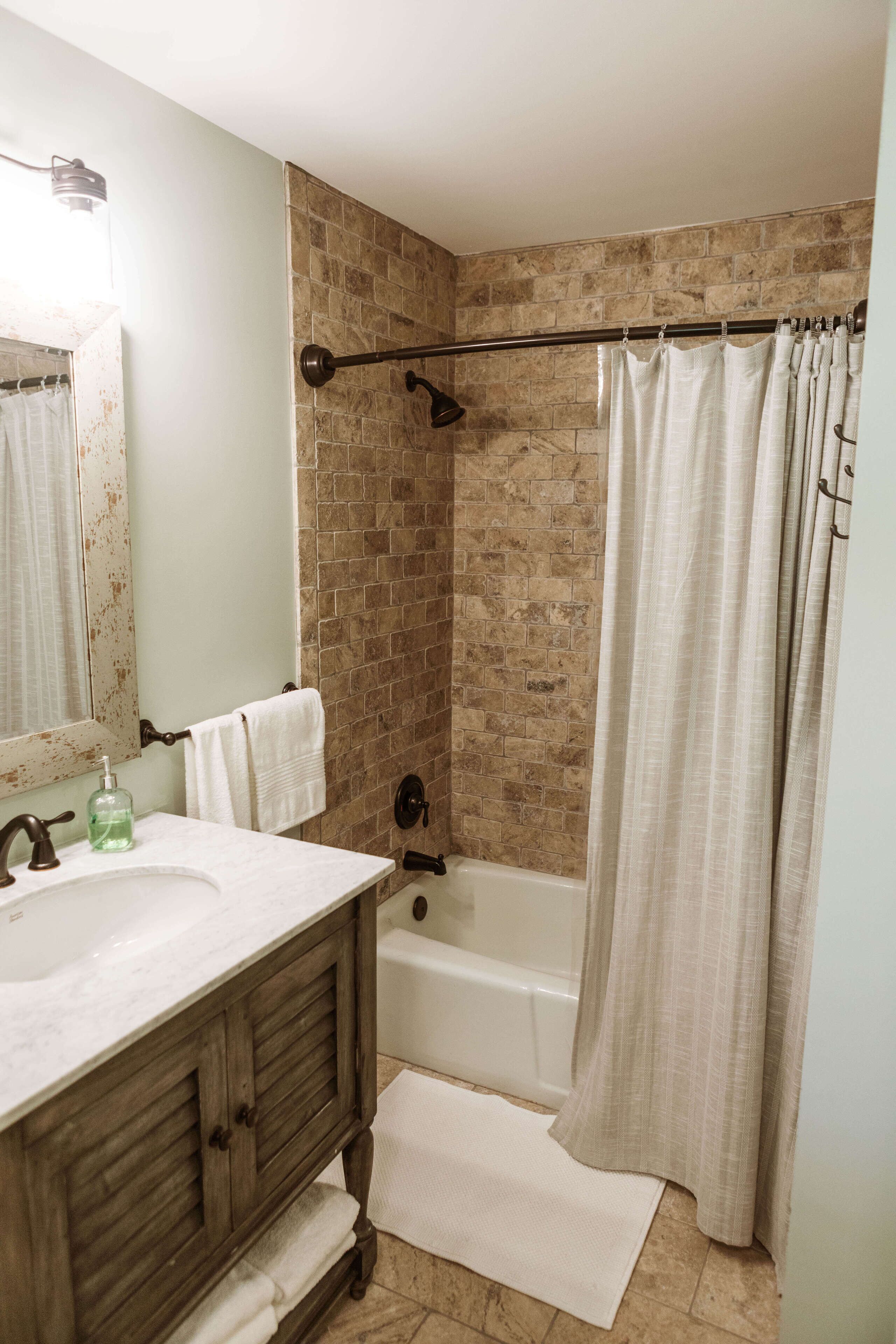Un baño con tocador de madera, un lavabo blanco, un espejo, una ducha con cortina color beige y una pared de ladrillo.
