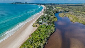 An aerial view of a coastal area with a beach, a river, and a cluster of buildings surrounded by greenery.