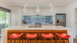 Une cuisine moderne dotée d’un îlot central, d’appareils électroménagers en acier inoxydable et d’une grande fenêtre avec des stores.