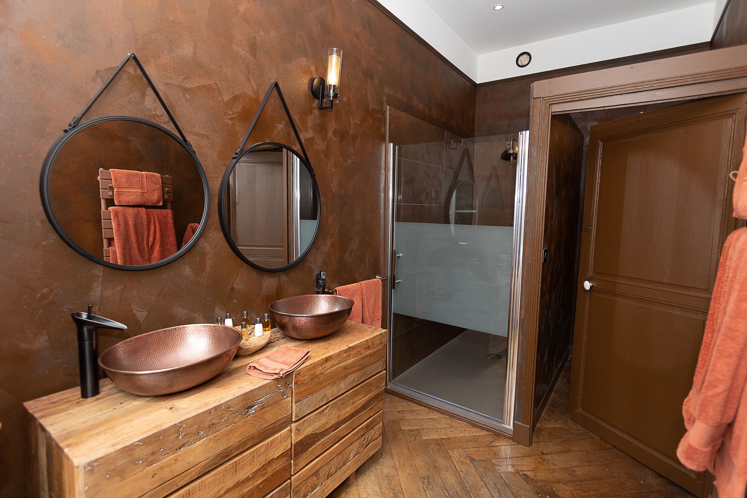 Ein Badezimmer mit einem Holzwaschtisch, zwei runden Spiegeln, einem Kupferbecken und einer Duschkabine aus Glas.