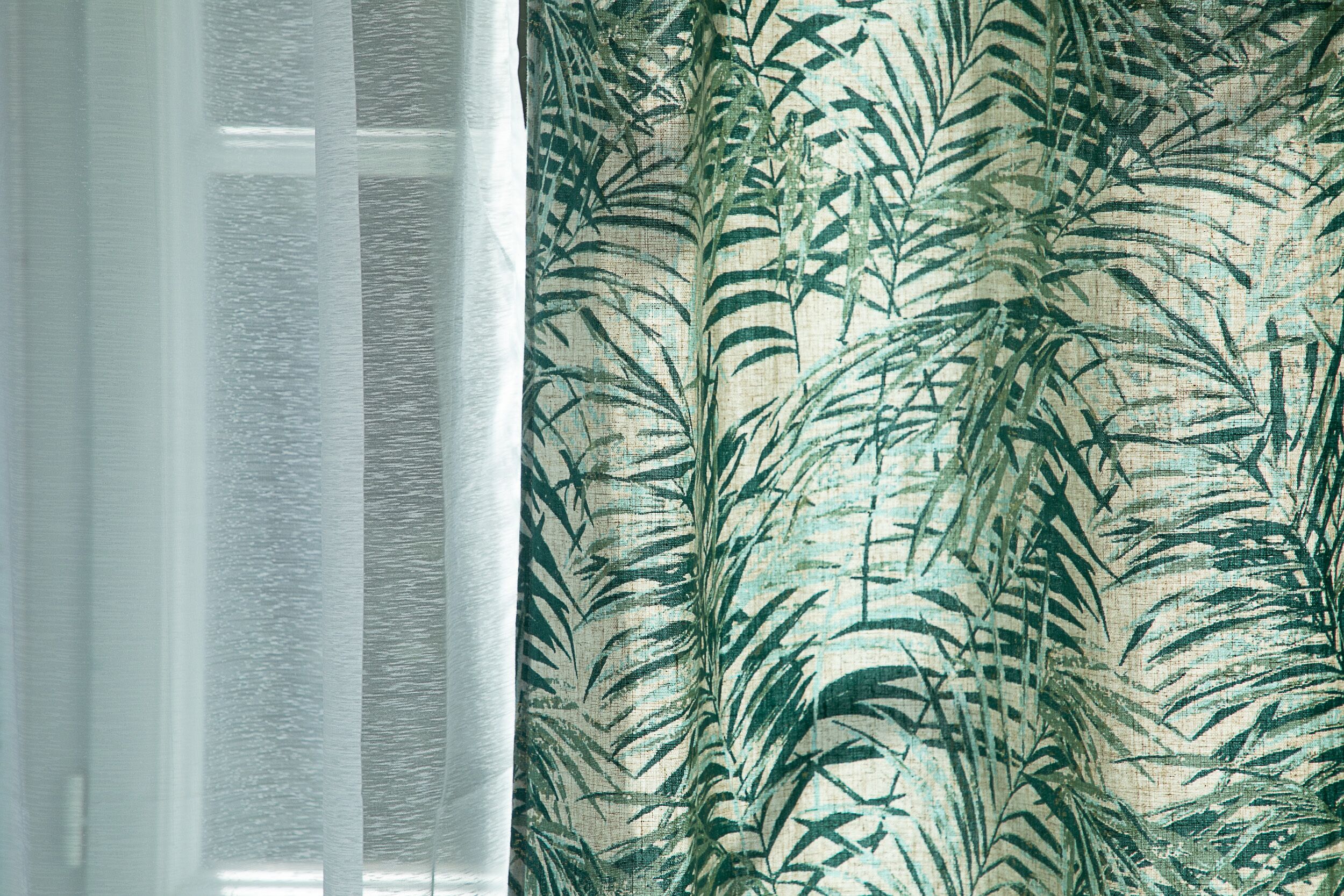 A window with sheer white curtains and a patterned fabric with palm leaves.