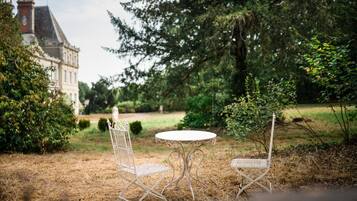 A white metal table and two chairs in a garden with a large tree and a building in the background.