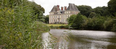 A large, historic building with multiple chimneys and a symmetrical facade, surrounded by lush greenery and a calm body of water.