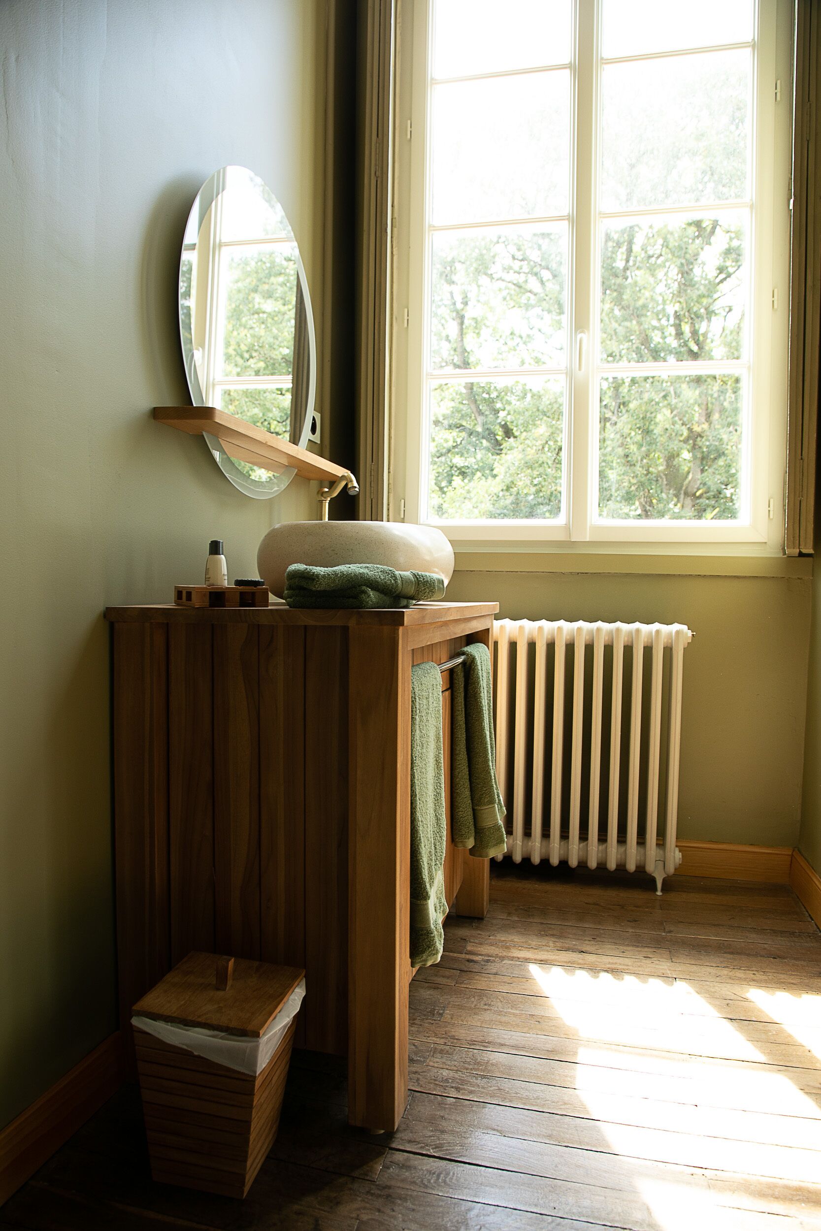A bathroom with a wooden vanity, a mirror, a sink, a towel, a radiator, and a window with a view of trees.