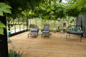 Um deck de madeira com área de estar ao ar livre, uma pérgula com parreiras de uva e vista para um jardim.