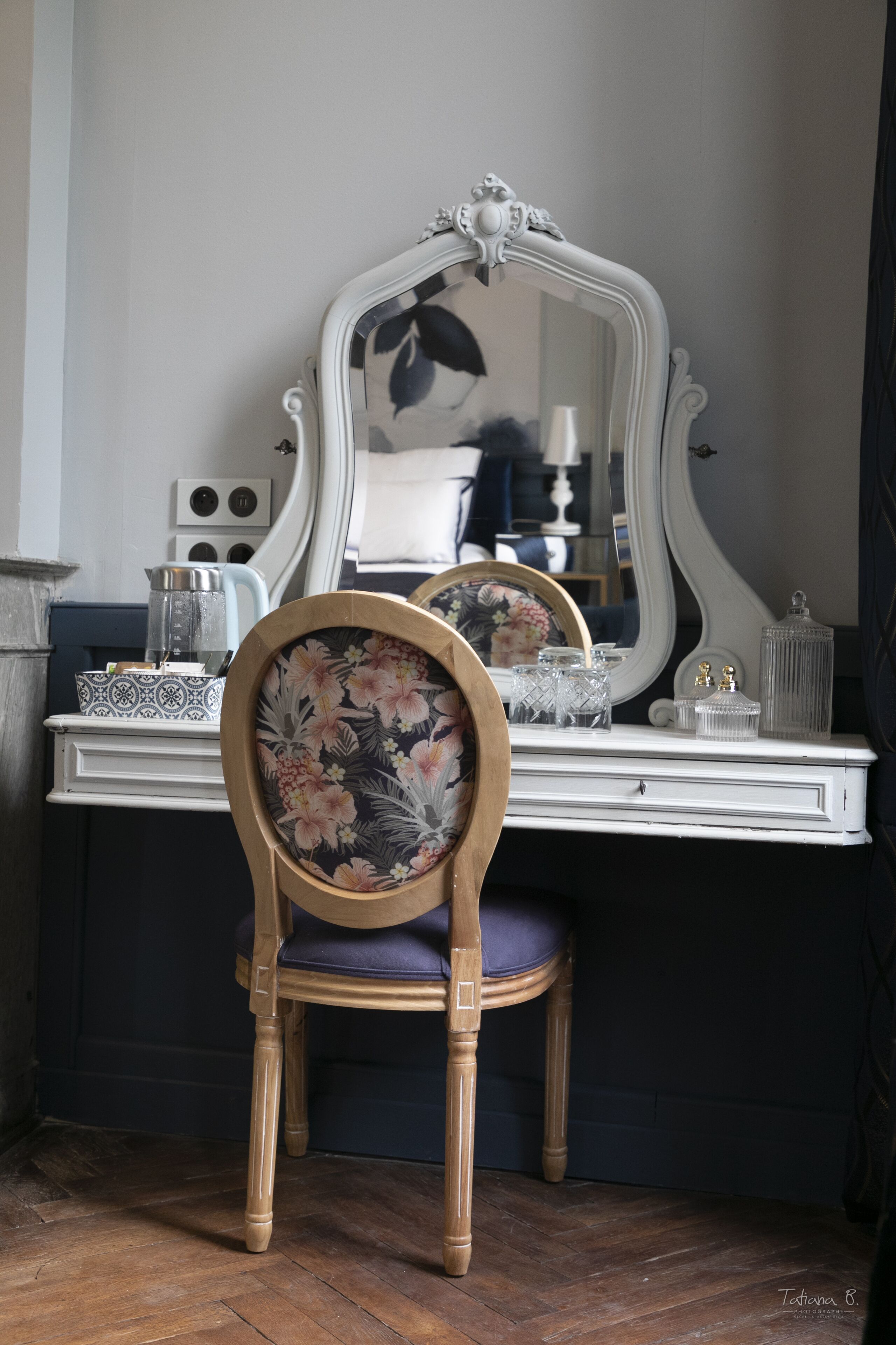 Une chaise en bois recouverte d’un tissu à motifs floraux, placée devant une commode blanche ornée d’un miroir, sur laquelle se trouvent une bouilloire et divers objets décoratifs.