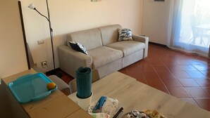 Un salon avec un canapé beige, une table basse sur laquelle se trouvent divers objets, un plateau bleu et une lampe. Il y a trois tableaux accrochés au mur et une fenêtre avec un rideau.