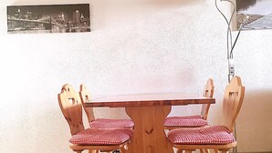 Une table à manger entourée de quatre chaises en bois, chacune agrémentée d’un coussin à carreaux. Un tableau représentant un paysage urbain en noir et blanc est accroché au mur derrière la table.