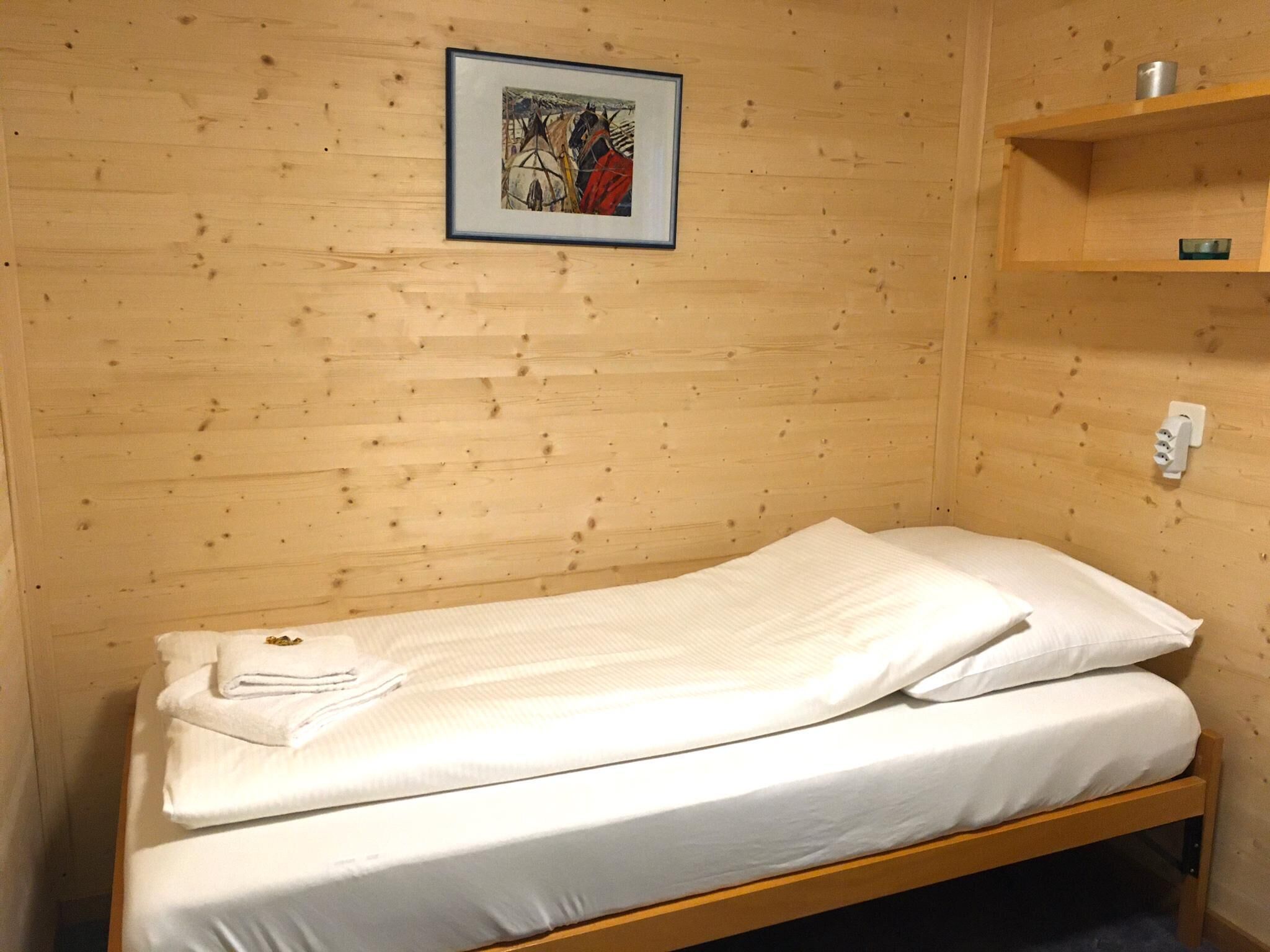 Ein Einzelbett mit weißen Laken in einem Holzraum, an der Wand hängt ein gerahmtes Bild.