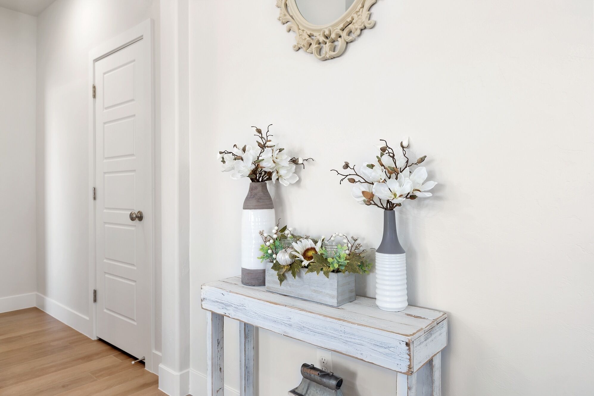 A hallway with a white door, a rustic wooden console table with two vases and a small plant, and a decorative mirror on the wall.