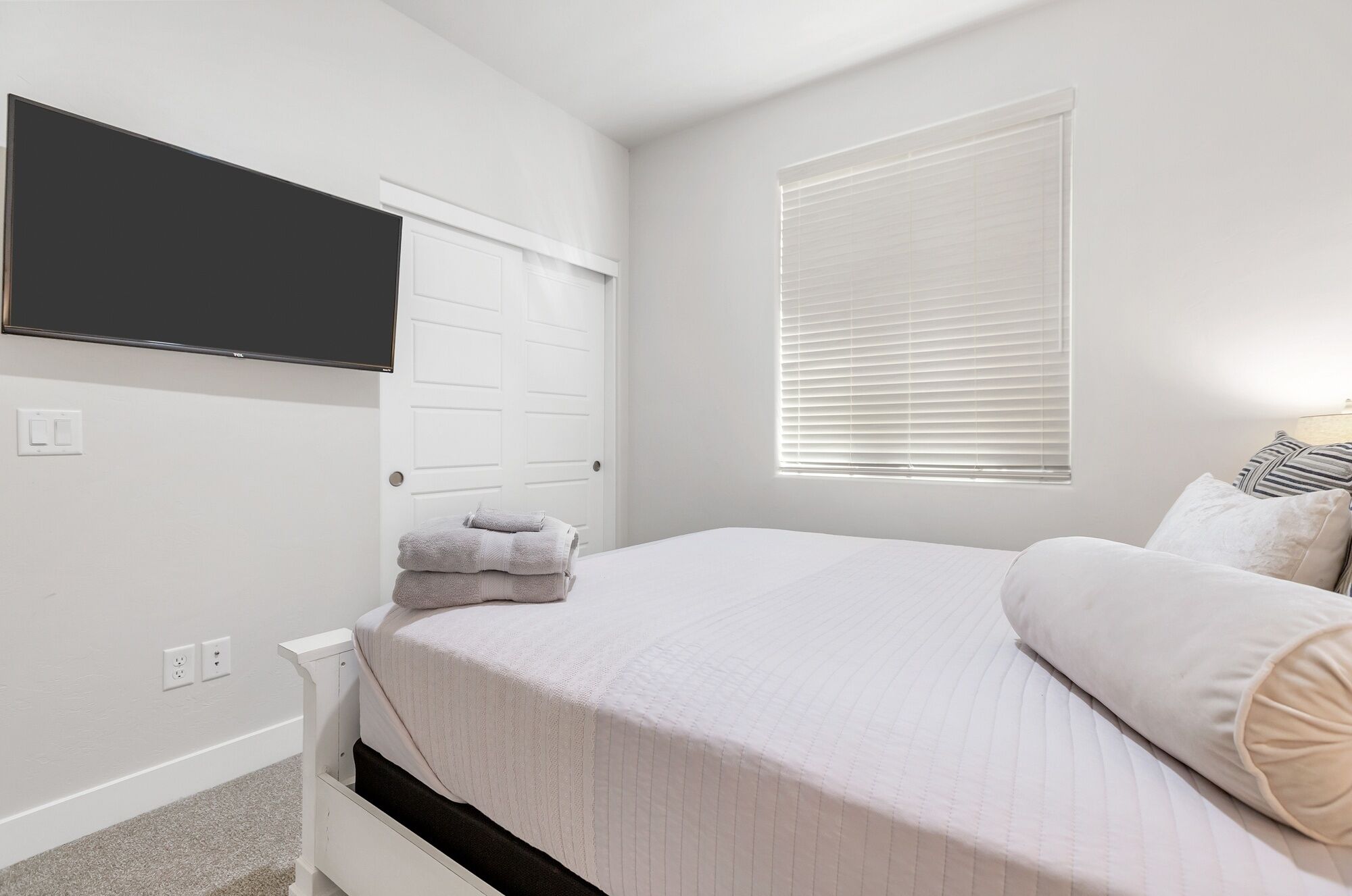 A bedroom with a bed, a TV mounted on the wall, a window with blinds, and a door.