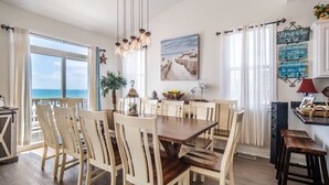 A dining area with a wooden table and chairs, a view of the sea through large windows, and a decorative wall sign.