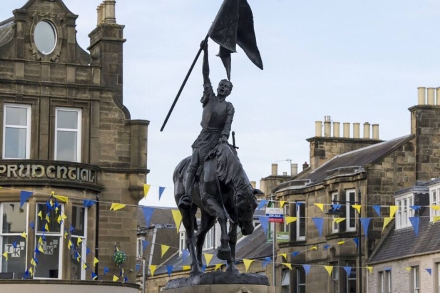 A statue of a figure on horseback with a flag, set against a backdrop of historic buildings and a sign reading "PRUDENTIAL".
