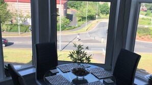 Zona pranzo con tavolo apparecchiato per quattro, circondato da sedie, e vista su strada e vegetazione attraverso una finestra.