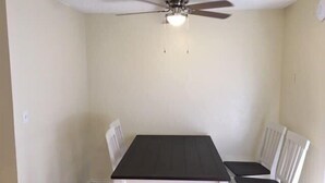 Un coin repas avec une table en bois foncé et des chaises blanches, un ventilateur de plafond et un luminaire.