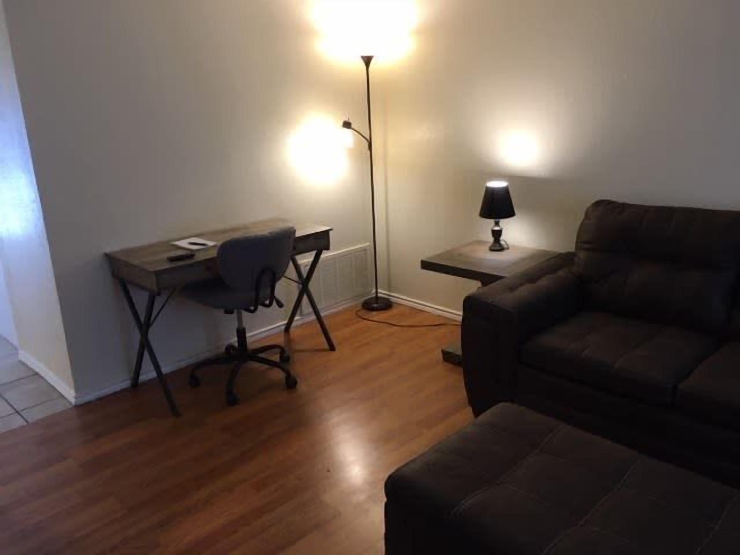 Una habitación con piso de madera, un escritorio con una silla, una lámpara y un sofá marrón.