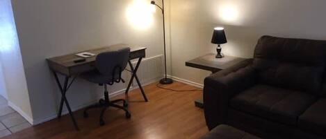 Una habitación con piso de madera, un escritorio con una silla, una lámpara y un sofá marrón.