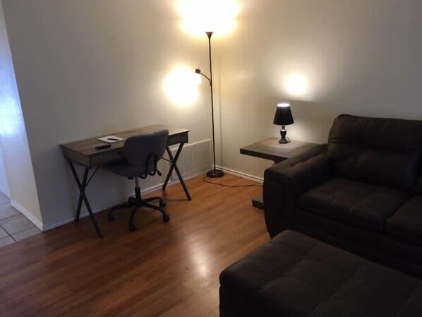 Una habitación con piso de madera, un escritorio con una silla, una lámpara y un sofá marrón.