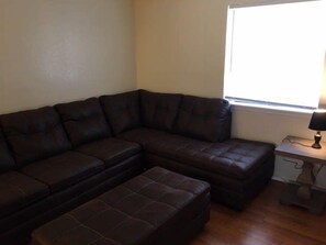 Una sala de estar con un sofá seccional marrón oscuro, un reposapiés a juego y una mesita de noche con una lámpara.