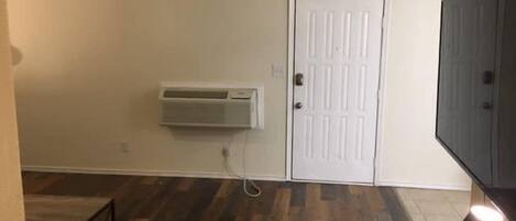 A room with a white door, an air conditioning unit mounted on the wall, a wooden floor, and a table with a lamp.