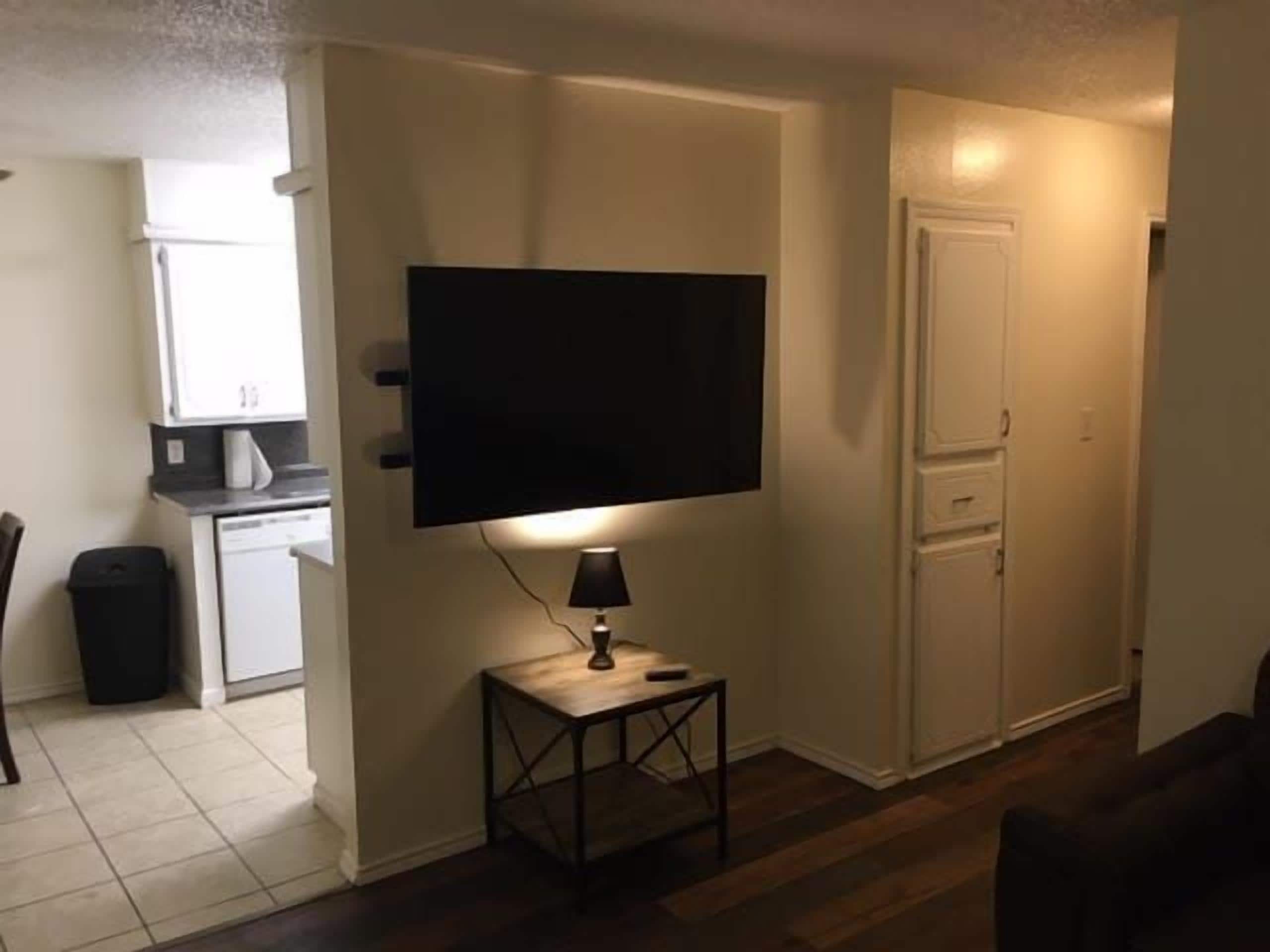 A compact living space with a flat-screen TV mounted on the wall, a small table with a lamp, and a kitchen area in the background.