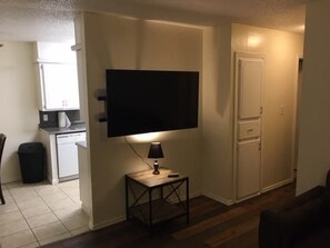 Un espacio habitable compacto con un televisor de pantalla plana montado en la pared, una mesita con una lámpara y una cocina al fondo.